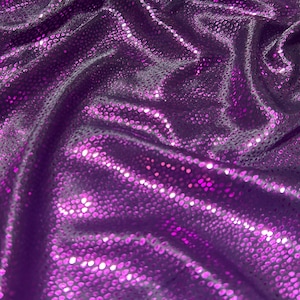 Op de afbeelding: Close-up van een glanzende, getextureerde stof in paarse tinten. Het materiaal heeft een subtiel, gestippeld patroon en is gedrapeerd in zachte plooien, waardoor highlights en schaduwen ontstaan. De stof lijkt een decoratief textiel te zijn.