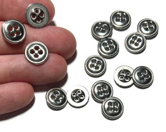 May include: A selection of small, silver buttons with a four-hole pattern. The buttons are circular with a slightly raised rim and a polished finish. Some are held in a hand, while others are scattered on a white surface.