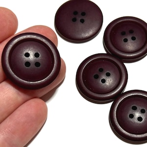 May include: A collection of dark burgundy buttons with four holes each. The buttons are round and have a slightly raised edge. One button is held in a hand, while the others are scattered on a white surface.