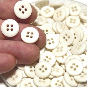 Può includere: Una collezione di bottoni rotondi color avorio, ciascuno con quattro fori. Alcuni bottoni sono tenuti in mano, gli altri in un contenitore trasparente. I bottoni sono realizzati in un materiale liscio e opaco, adatti per cucito e artigianato.