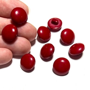 May include: A collection of glossy, deep red buttons in various sizes and shapes. The buttons are oval and round, with a smooth, reflective surface. One button has a shank on the back. The buttons are scattered on a white background.