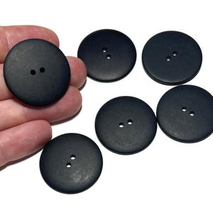 May include: A collection of six large, round, matte black buttons. Each button has two small holes for sewing. The buttons are made of a solid material and are suitable for various sewing projects.