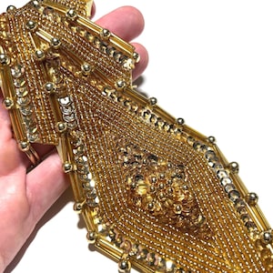 May include: Gold beaded trim with sequins and small gold beads. The trim has a geometric design with a central diamond shape. The trim is held in a hand against a white background. This trim is suitable for embellishing clothing or accessories.