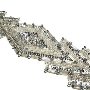 May include: Silver beaded trim with a geometric design. The trim features a central diamond shape with intricate sequin detailing, surrounded by rows of small beads and larger silver accents. Ideal for embellishing garments or accessories.