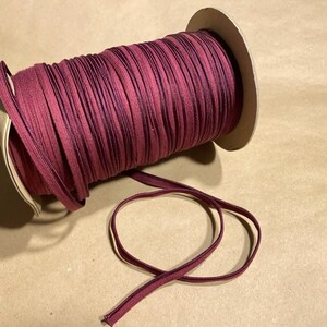 May include: A spool of burgundy elastic ribbon. The ribbon is tightly wound around a cardboard core. A length of the ribbon extends from the spool, showing a dark purple edge. The ribbon is likely used for sewing or crafting projects.