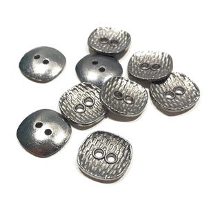 Peut inclure: Un ensemble de boutons argentés de forme carrée, chacun avec deux trous. Certains ont une surface texturée, d'autres sont lisses. Les boutons sont dispersés sur un fond blanc.