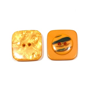May include: Two square buttons, one with a marbled gold finish and two holes, the other with a solid orange finish and a black and orange striped button inside.