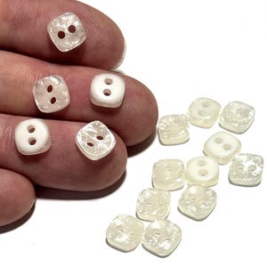 May include: A collection of square, two-hole buttons in varying shades of white and off-white. Some buttons are opaque, while others are translucent, with a textured surface. The buttons are shown against a white background.