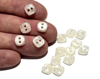 May include: A collection of square, two-hole buttons in varying shades of white and off-white. Some buttons are opaque, while others are translucent, with a textured surface. The buttons are shown against a white background.