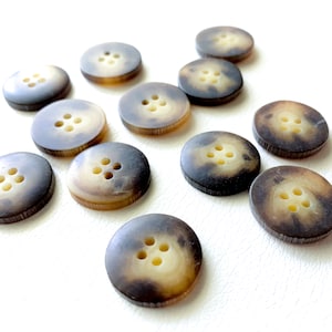 Puede incluir: Una colección de diez botones de color marrón y crema con cuatro agujeros cada uno. Los botones están hechos de un material natural, posiblemente cuerno o hueso.