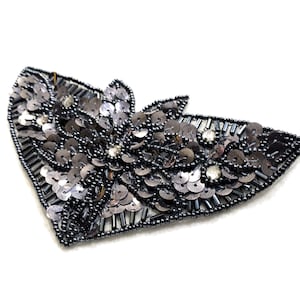 May include: A silver and black sequined applique in the shape of a stylized butterfly. The applique is made of small, round sequins and beads.