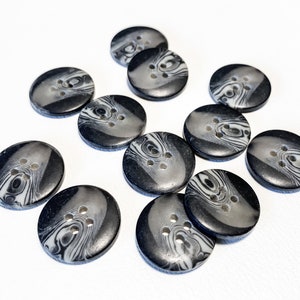 May include: A collection of marbled-effect buttons in shades of black and gray. Each button has a swirl pattern and four holes for sewing. The buttons are round and oval, scattered on a white surface.