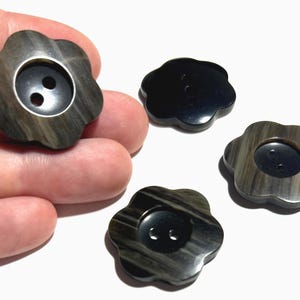 May include: A collection of flower-shaped buttons. The buttons have a dark, swirling pattern with a black center and two holes for sewing. One button is held in a hand, with three others scattered on a white surface.