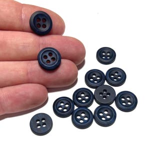 May include: A collection of navy blue buttons with four holes each. Some buttons are held in a hand, while others are scattered on a white surface. The buttons have a slightly textured appearance and a matte finish. Ideal for sewing and crafts.