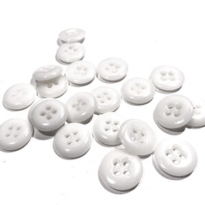 May include: A collection of white plastic buttons with four holes each. The buttons are scattered on a white surface.
