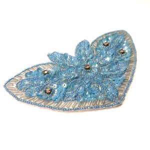 May include: A light blue, sequined applique in a floral design. The applique is shaped like a leaf, with a beaded silver border. The floral design is embellished with small, round, iridescent sequins and small, round, brown rhinestones.