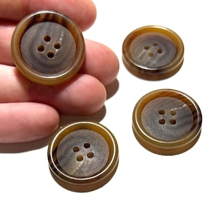 May include: Four round, brown buttons with four holes each. The buttons have a light brown outer ring and a darker brown center with a wood grain pattern. One button is held in a hand.