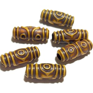 Puede incluir: Seis cuentas en forma de barril, marrones y amarillas, con intrincados diseños tallados. Las cuentas tienen un motivo central en forma de ojo y están rodeadas de bandas acanaladas. Las cuentas están dispuestas sobre un fondo blanco.