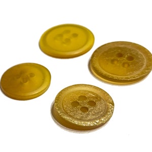 May include: Four yellow buttons with four holes each are arranged on a white surface. The buttons vary in size and have a slightly textured appearance. The buttons are a warm, golden yellow color.
