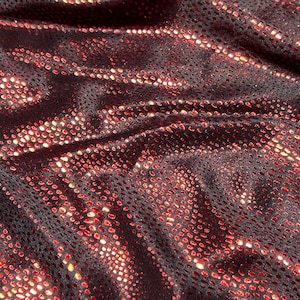 Op de afbeelding: Close-up van een zwarte stof met een rood, gestippeld patroon. De stof is gedrapeerd, waardoor schaduwen en highlights ontstaan die de textuur en glans benadrukken. Het materiaal lijkt flexibel en mogelijk gebruikt voor kleding.