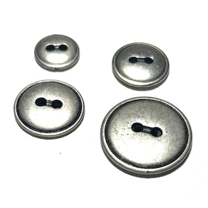 May include: Four silver-toned buttons of varying sizes are arranged on a white surface. Each button has a two-hole design. The buttons have a slightly aged, metallic appearance, suggesting a vintage or antique style, suitable for sewing or crafts.