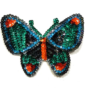 May include: A green, blue, and orange sequined butterfly applique. The butterfly is made of small, iridescent sequins that are sewn together to create a detailed design.