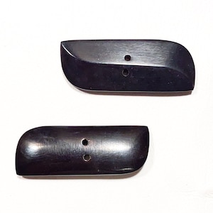 May include: Two black horn buttons with two holes each. The buttons are oval shaped with rounded edges.