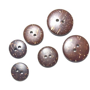 May include: A collection of six round, brown coconut buttons of varying sizes, each with two holes for sewing. The buttons have a natural, textured appearance with lighter brown and tan markings, set against a bright white background.