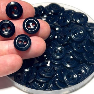 May include: A collection of navy blue buttons with two holes, some held in a hand, and the rest in a clear plastic container. The buttons are round and have a glossy finish, suitable for sewing or crafts.