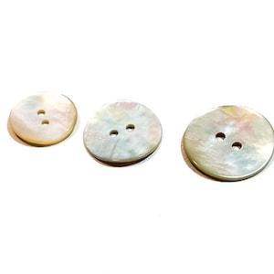 May include: Three round, iridescent mother-of-pearl buttons with two or three holes each. The buttons vary slightly in size and have a pearlescent sheen with hints of pink, green, and yellow. They are set against a white background.