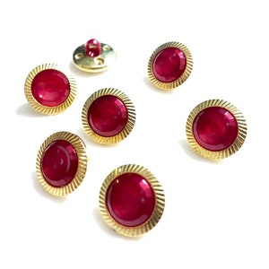 May include: Seven red and gold buttons with a decorative, ridged edge. The buttons have a shiny, metallic gold finish and a deep red center.