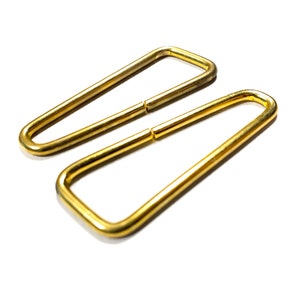 May include: Two gold-colored metal D-rings, one slightly larger than the other, are shown on a white background.