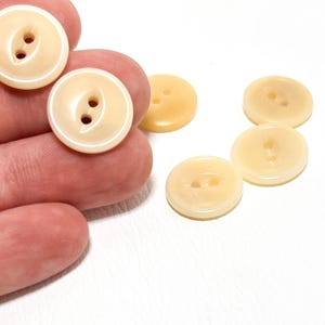 May include: A collection of round, cream-colored buttons with two holes each. Some buttons are held in a hand, while others are scattered on a white surface. The buttons have a glossy finish and a slightly off-white hue.