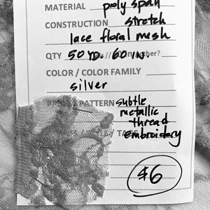 May include: A white tag with black text listing the details of a silver lace fabric. The tag reads "ITEM # F0396", "MATERIAL poly span stretch lace floral mesh", "QTY 50 YDs/60 IN", "COLOR/COLOR FAMILY silver", "PATTERN subtle metallic thread embroidery", and "46".