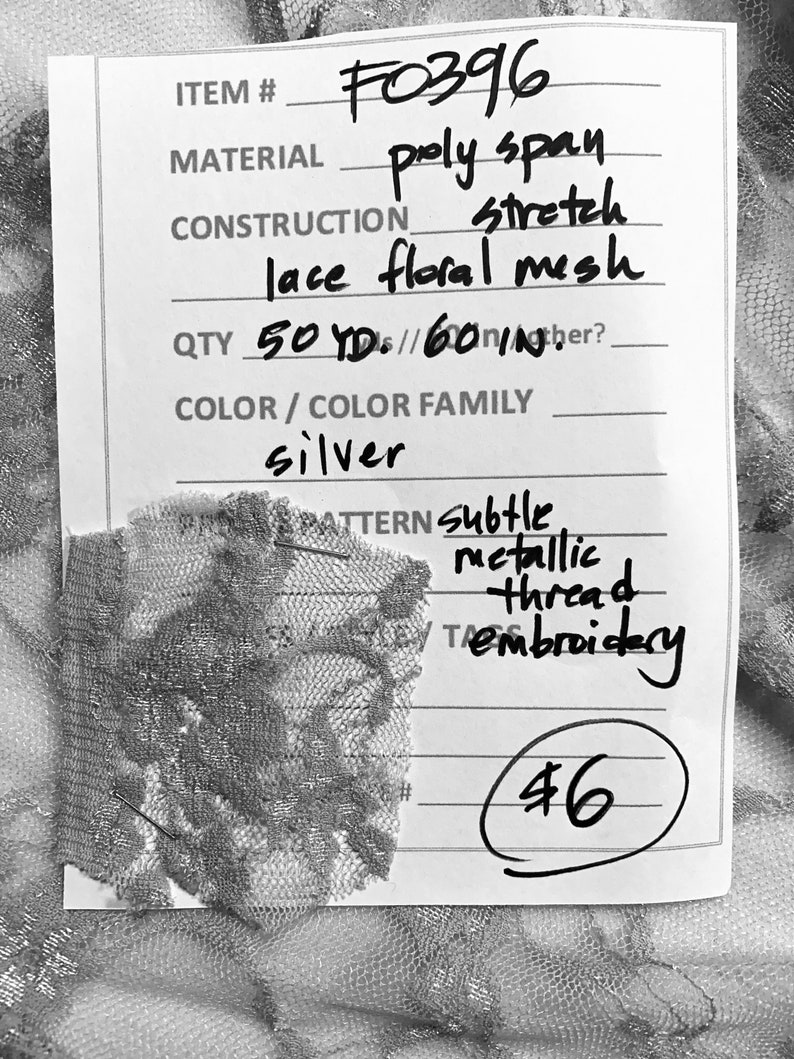 May include: A white tag with black text listing the details of a silver lace fabric. The tag reads "ITEM # F0396", "MATERIAL poly span stretch lace floral mesh", "QTY 50 YDs/60 IN", "COLOR/COLOR FAMILY silver", "PATTERN subtle metallic thread embroidery", and "46".