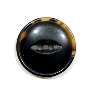 May include: A black oval button with a brown and yellow tortoiseshell rim. The button has two holes and a decorative groove.
