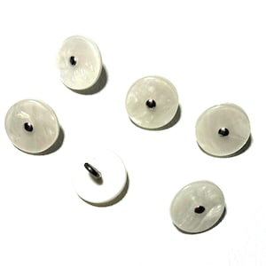 May include: Six off-white, round buttons with a pearlescent finish. Each button features a small, dark metal shank and a matching dark metal centre. The buttons are scattered on a white surface.