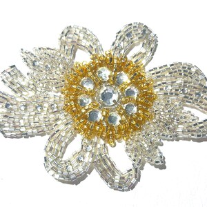 May include: A decorative beaded flower applique. The central part is gold, surrounded by silver petals. Clear rhinestones are set in the centre. The applique has a bow-like design, suitable for clothing or accessory embellishment.