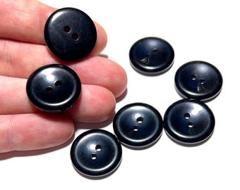 May include: A collection of seven glossy, black buttons with two holes each. The buttons are round and have a slightly domed shape. They are scattered on a white surface, with some held in a hand.
