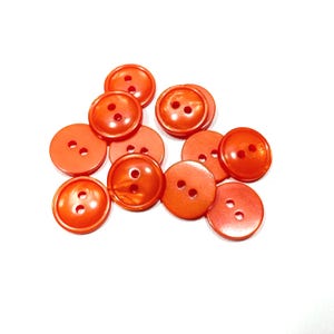 May include: A collection of orange, round buttons with two holes each. The buttons are scattered on a white surface. The buttons have a glossy finish and vary slightly in shade.