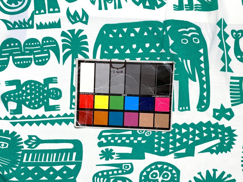 May include: White fabric with a repeating pattern of teal-colored stylized animals and plants. The design includes elephants, crocodiles, and other creatures. A color calibration card is in the foreground.
