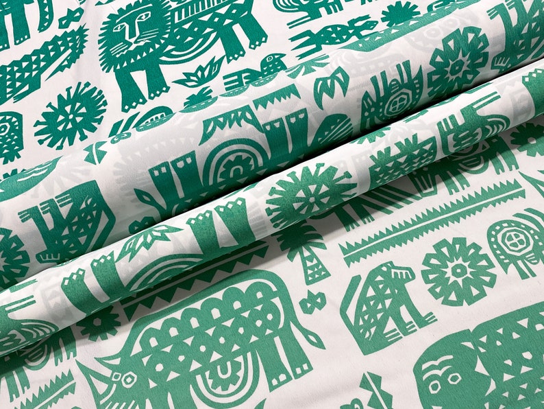 May include: White fabric with a repeating pattern of green stylized animals and plants. The design includes lions, elephants, birds, and palm trees. The fabric is folded, showing the pattern's repeat.