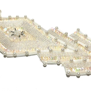 May include: A decorative, off-white beaded trim with a geometric design. The trim features iridescent sequins, small pearls, and silver-colored seed beads. The central design element is a diamond shape with a pearl and bead cluster.