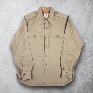 May include: A tan long-sleeve shirt with two chest pockets and button closures. The shirt has epaulets on the shoulders and a collar with a brown lining. The tag on the collar reads "SALUTE".