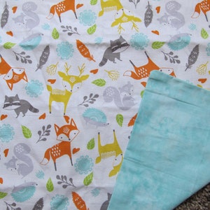 May include: A white fabric baby blanket with a woodland animal print featuring foxes, deer, raccoons, squirrels, and hedgehogs. The blanket has a light blue minky fabric on the back side.