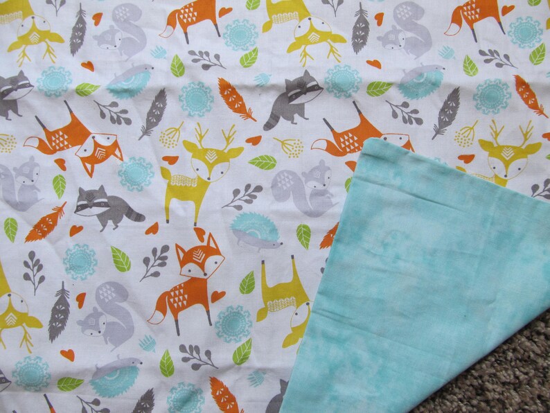 May include: A white fabric baby blanket with a woodland animal print featuring foxes, deer, raccoons, squirrels, and hedgehogs. The blanket has a light blue minky fabric on the back side.