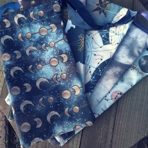 May include: Five fabric pouches with celestial designs. The pouches are made of a soft, blue fabric with gold accents. The designs include moons, stars, and constellations.