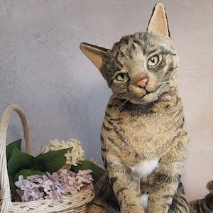 Puede incluir: Un juguete de peluche realista en forma de gato con marcas de tabby marrones y grises. El gato está sentado con las piernas cruzadas y la cola enroscada alrededor de su cuerpo. El gato tiene una mancha blanca en el pecho y ojos verdes.