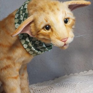 Realistic soft stuffed orange tabby Oriental breed cat toy has yellow taxidermy eyes, pink colored polymer clay paws, white translucent polymer clay claws, and artificial whiskers. The cat  has a green scarf with a diamond pattern. Perfect for collectors, gifts, memorial pet, and home decor.