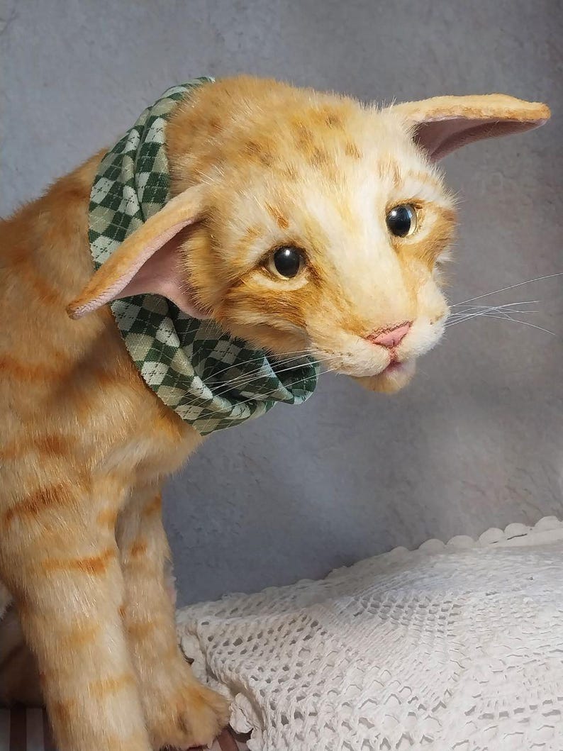 Realistic soft stuffed orange tabby Oriental breed cat toy has yellow taxidermy eyes, pink colored polymer clay paws, white translucent polymer clay claws, and artificial whiskers. The cat  has a green scarf with a diamond pattern. Perfect for collectors, gifts, memorial pet, and home decor.